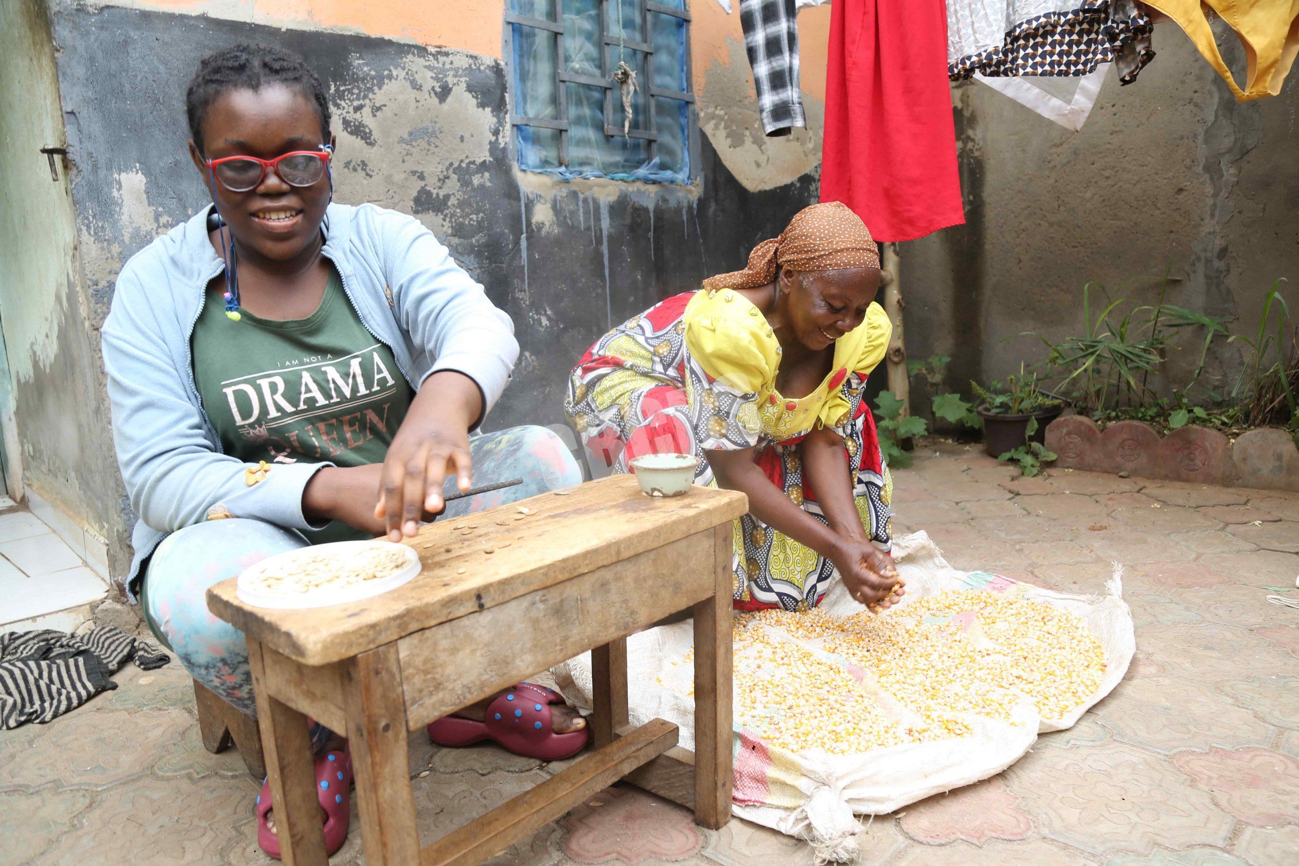Esthelle cracking egusi with focus and care while her mother gathers maize nearby—sharing moments of daily life and teamwork in front of their home.